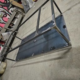 KLARIGA-WELD Maciej Burdziński - Stelaż z profili i blachy, prawdopodobnie do mebla, ze śladami spawania, leżący na betonowej posadzce w warsztacie, w tle drabina i ściana z graffiti.