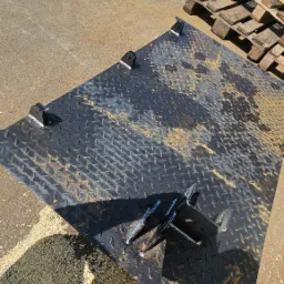 Metalowa płyta z wytłaczanym wzorem i przyspawanymi uchwytami, leżąca na ziemi z wiórami, obok palet.