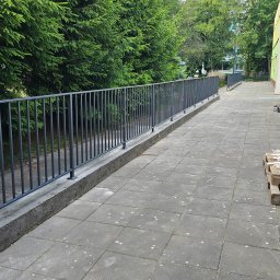 KLARIGA-WELD Maciej Burdziński - Długa, szara balustrada z pionowych prętów zabezpieczająca chodnik wzdłuż budynku, obok paleta z drewnianymi listwami.