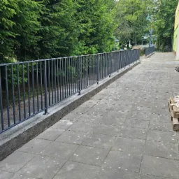 Długa, szara balustrada z pionowych pręt&oacute;w zabezpieczająca chodnik wzdłuż budynku, obok paleta z drewnianymi listwami.