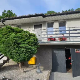 Dwukondygnacyjny dom z szarymi balustradami balkonowymi i schodowymi, żółtą skrzynką na listy i otwartymi drzwiami garażowymi.