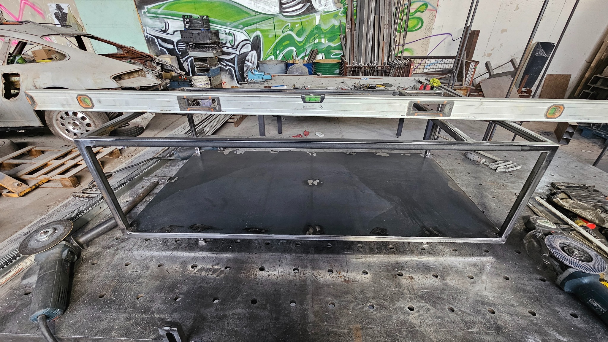 Metalowa rama z czarnym blatem na stole warsztatowym, obok szlifierka kątowa; w tle widoczny zniszczony samochód i graffiti na ścianie.