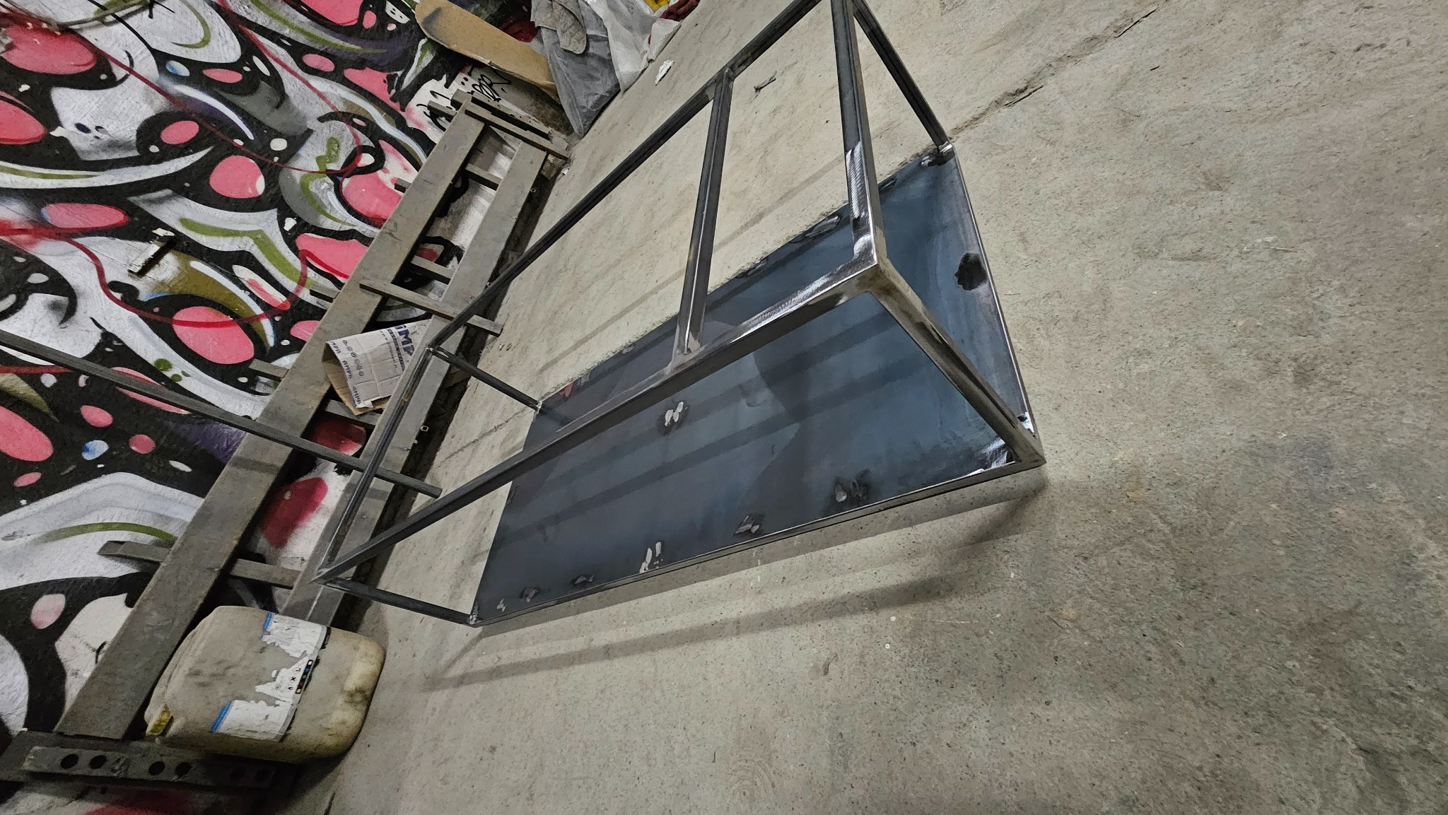 Stelaż z profili i blachy, prawdopodobnie do mebla, ze śladami spawania, leżący na betonowej posadzce w warsztacie, w tle drabina i ściana z graffiti.