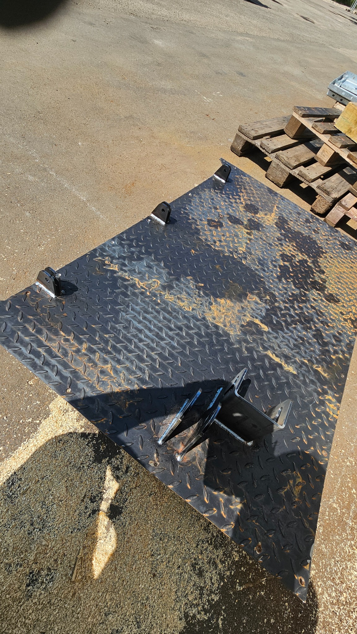 Metalowa płyta z wytłaczanym wzorem i przyspawanymi uchwytami, leżąca na ziemi z wiórami, obok palet.