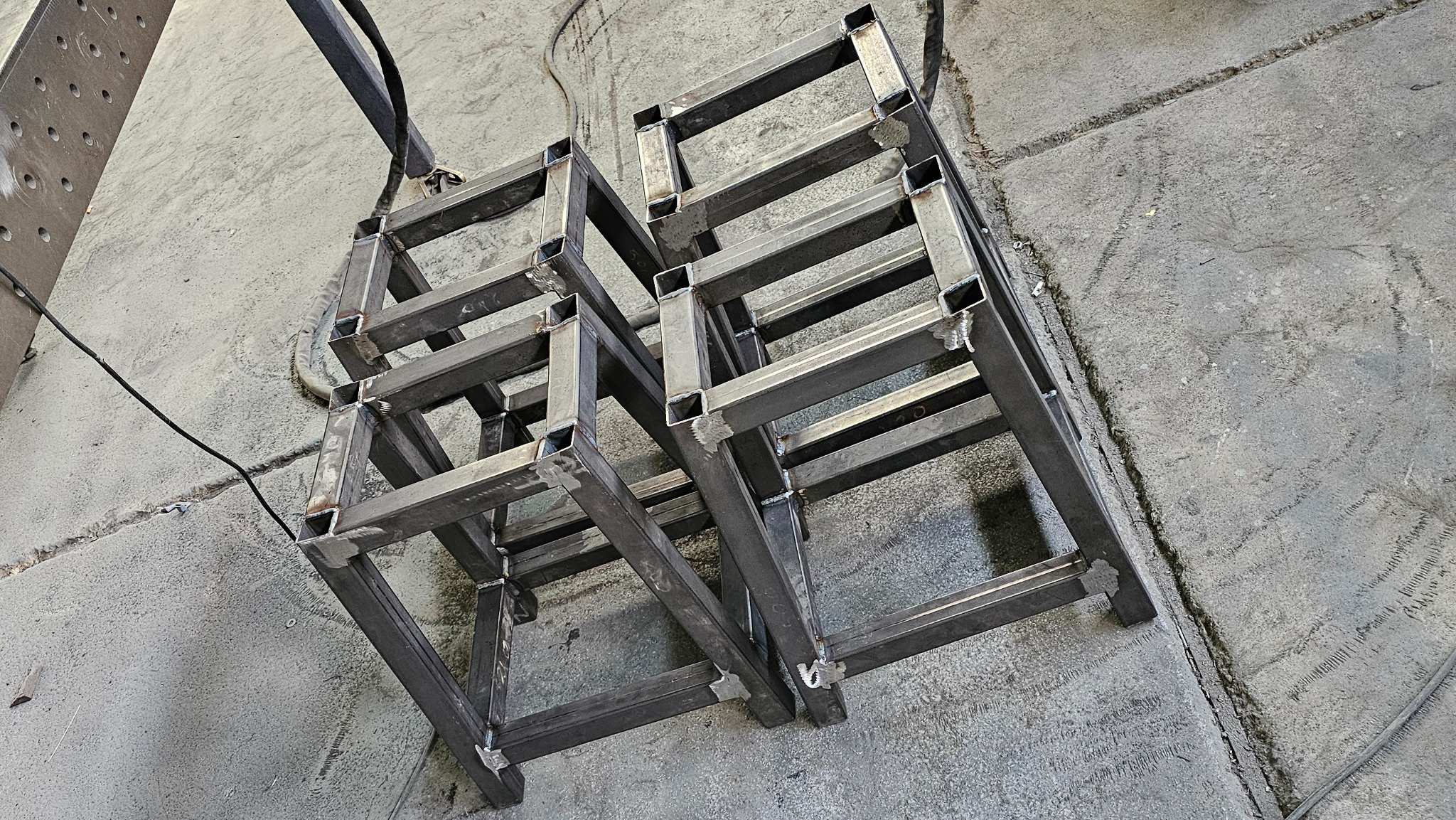 Surowe, pospawane metalowe konstrukcje w kształcie prostopadłościanów z kwadratowych profili na szarym betonowym podłożu, widoczne spawy na łączeniach.