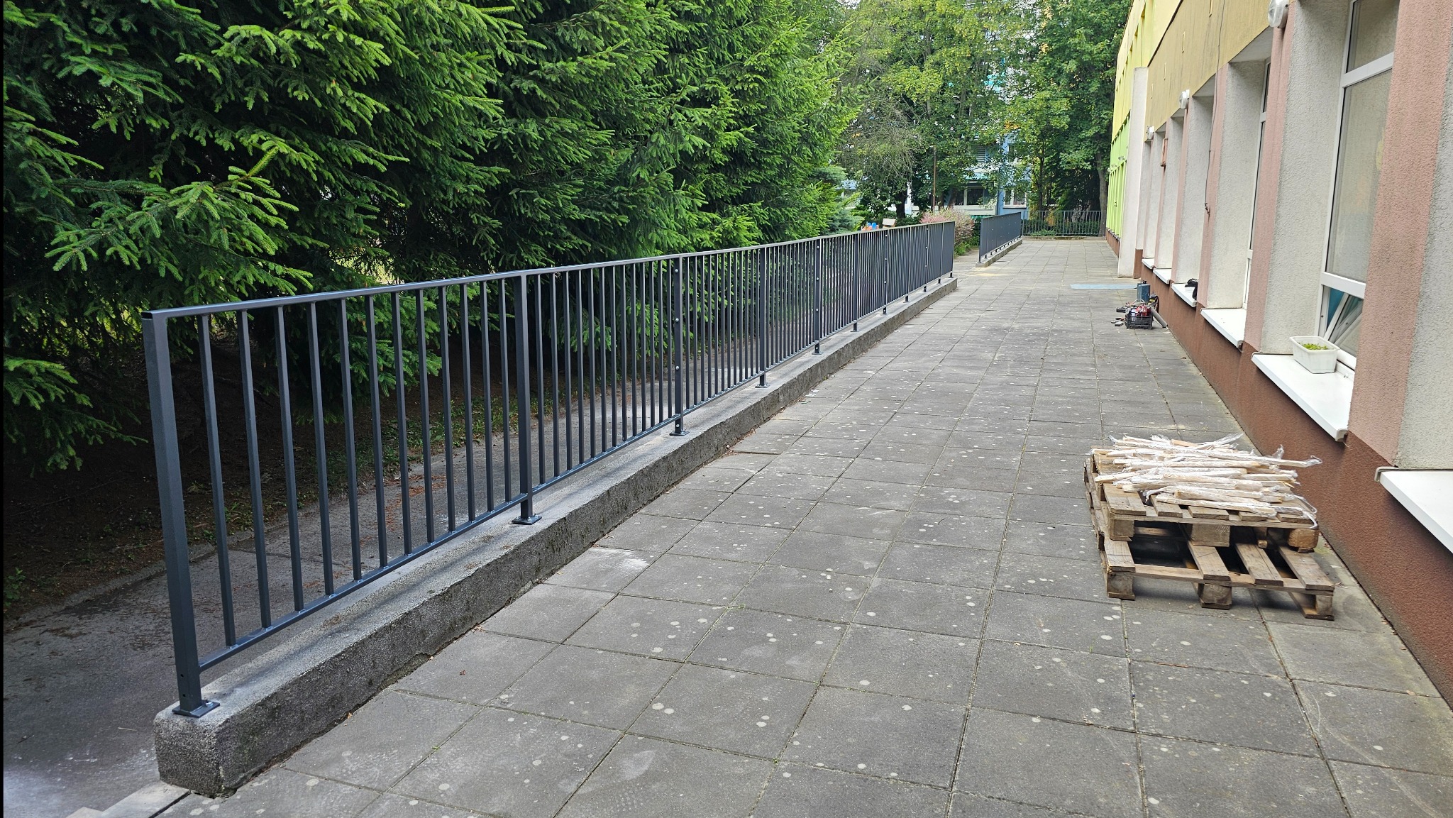 Długa, szara balustrada z pionowych prętów zabezpieczająca chodnik wzdłuż budynku, obok paleta z drewnianymi listwami.
