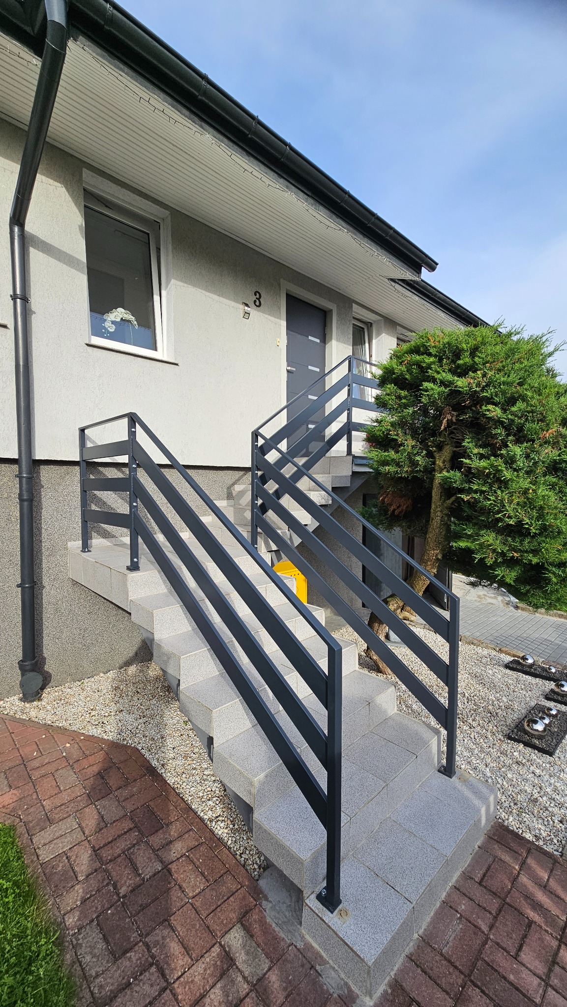 Zewnętrzne schody wejściowe z szarymi, prostymi poręczami z metalowych profili, prowadzące do drzwi budynku z numerem 3.