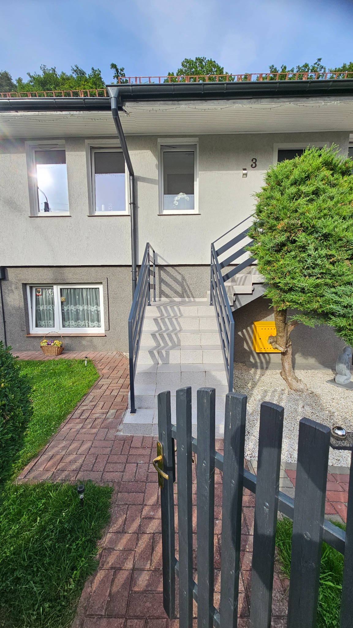 Zewnętrzne schody z szarymi metalowymi poręczami prowadzące do domu z numerem 3, widoczne zza drewnianego płotu z złotą klamką.