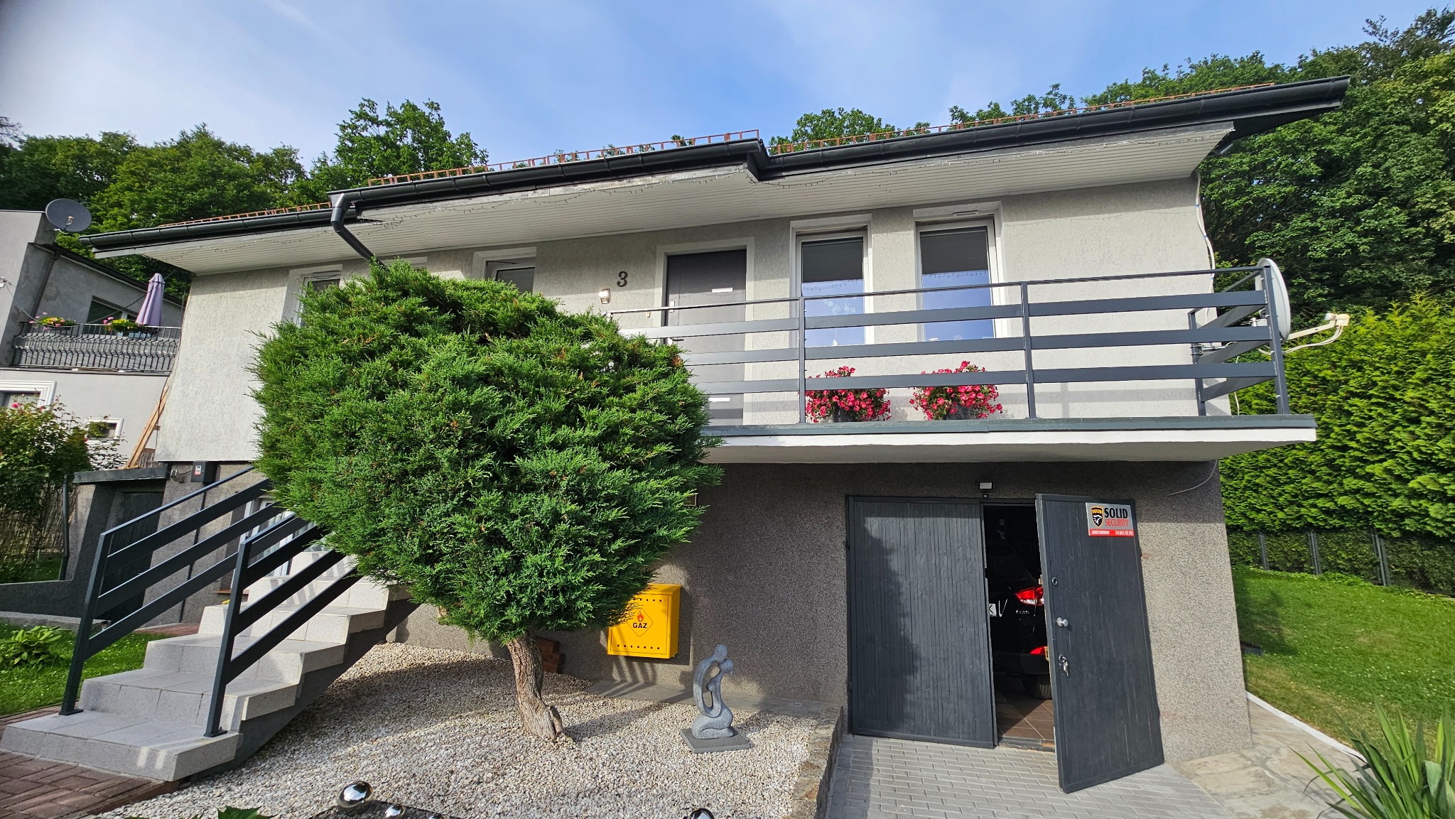 Dwukondygnacyjny dom z szarymi balustradami balkonowymi i schodowymi, żółtą skrzynką na listy i otwartymi drzwiami garażowymi.