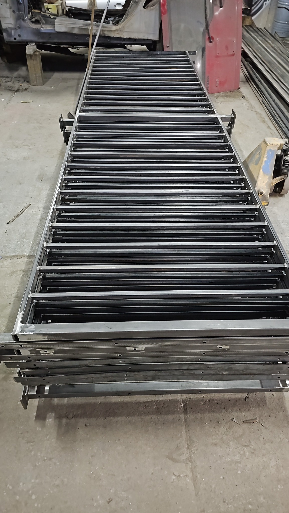 Stos metalowych konstrukcji kratowych z prostokątnymi otworami, leżących na sobie w warsztacie, widoczne elementy karoserii samochodowej w tle.