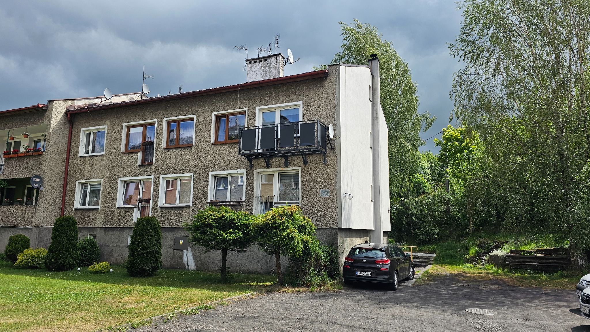 Szary blok z balkonem z czarną metalową balustradą, widok z zewnątrz w pochmurny dzień.