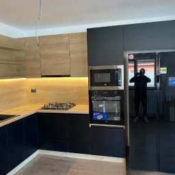 Meble - Nowoczesna kuchnia z drewnianymi i czarnymi frontami, podświetleniem LED, sprzętem AGD marki Beko i odbiciem fotografa w czarnej lodówce.