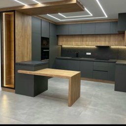 Meble - Nowoczesna kuchnia z szarymi szafkami, drewnianymi akcentami i wyspą z blatem z jasnego drewna, oświetlenie LED pod szafkami i na suficie.