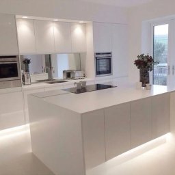 Meble - Minimalistyczna, biała kuchnia z wyspą, podświetleniem LED pod szafkami i dużym oknem balkonowym w tle.