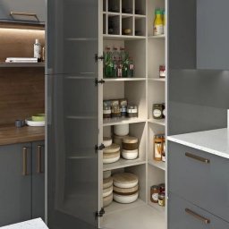 Meble - Szara, nowoczesna kuchnia z otwartą szafką narożną, wypełnioną słoikami, butelkami i pojemnikami, widoczne blaty z jasnego kamienia.