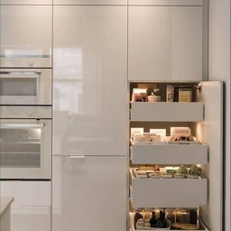 Meble - Biała zabudowa kuchenna z wbudowanymi piekarnikami i otwartą szafką cargo z wewnętrznym oświetleniem LED, ukazująca zawartość szuflad: przyprawy, przetwory i opakowania produktów spożywczych.