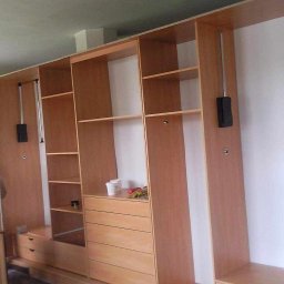Meble - Montaż modułowej szafy na wymiar z jasnego drewna, widoczne wysuwane wieszaki, półki i szuflady, pracownik w tle.