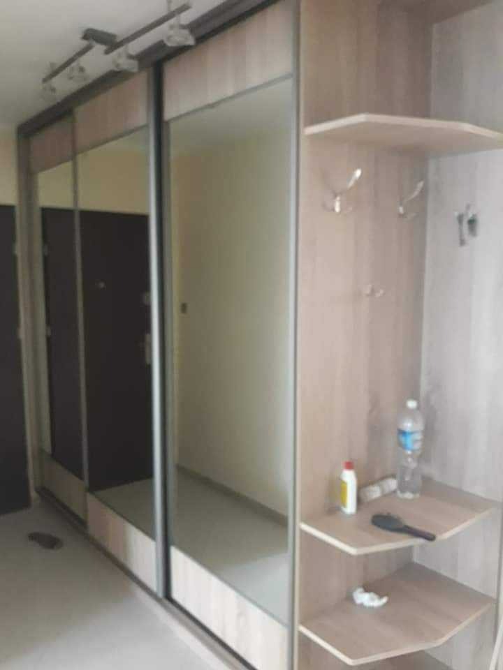 Jasna szafa przesuwna z lustrami i otwartym regałem na drobne przedmioty, widoczne haczyki na ubrania i półki z kosmetykami oraz butelką wody.