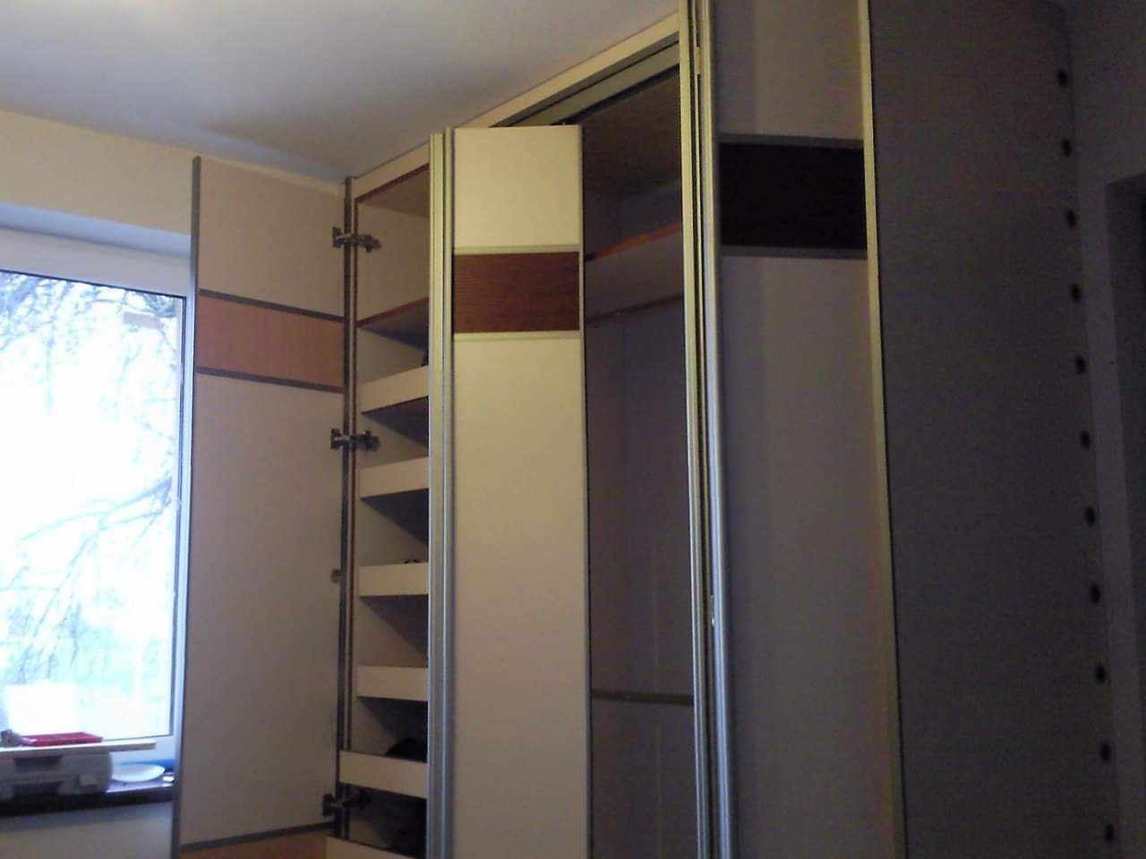 Otwarta szafa wnękowa z przesuwnymi drzwiami i półkami, widoczne elementy drewniane i metalowe mocowania, na tle okno z widokiem na drzewo.