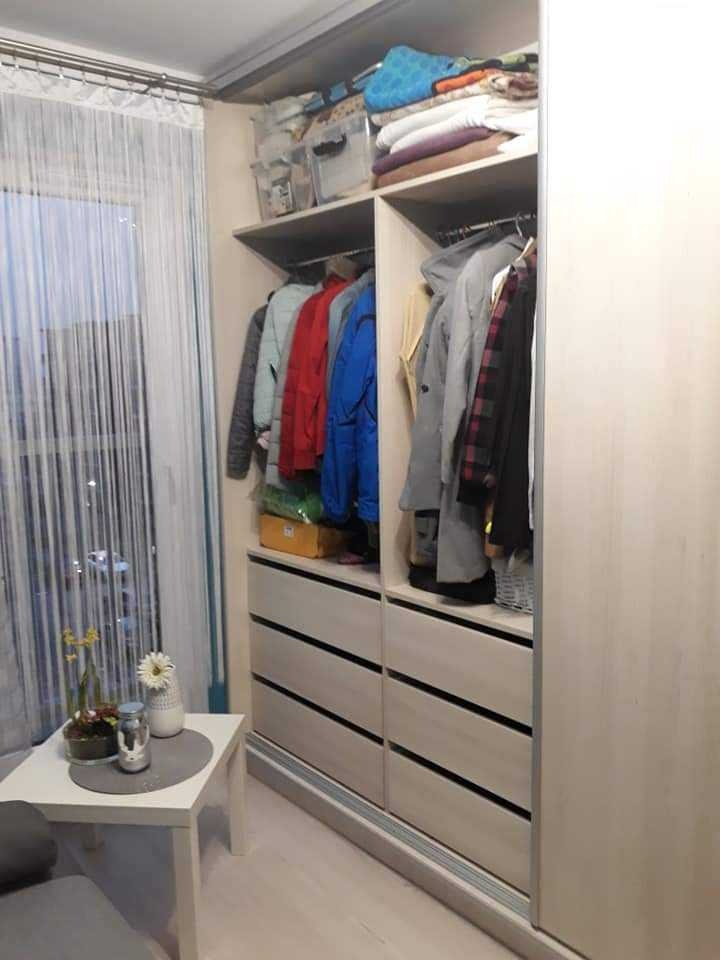 Wnętrze szafy przesuwnej z jasnego drewna, widoczne ubrania na wieszakach i na półkach, obok stolik kawowy z dekoracjami, firanka w oknie.