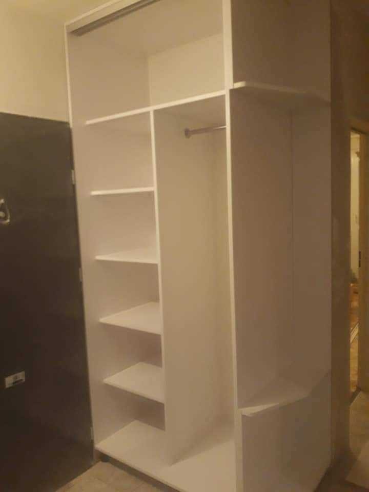 Wnętrze szafy na wymiar z białymi półkami i drążkiem na ubrania, widoczne z perspektywy pod kątem, z ciemnymi drzwiami w tle.