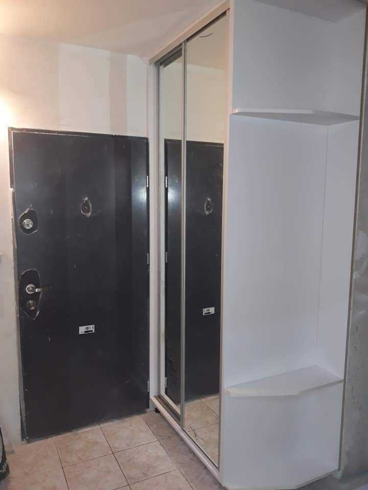 Biała szafa przesuwna z lustrami i półkami obok czarnych drzwi wejściowych w korytarzu z jasnymi kafelkami na podłodze.