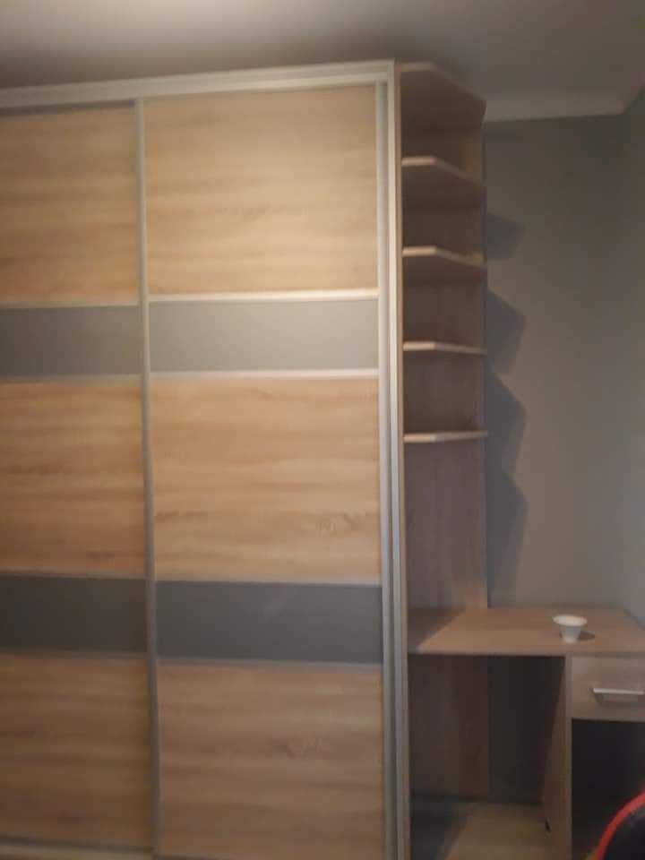 Drewniana szafa przesuwna z szarymi wstawkami, zintegrowana z biurkiem i półkami narożnymi, ustawiona w pokoju o szarych ścianach.