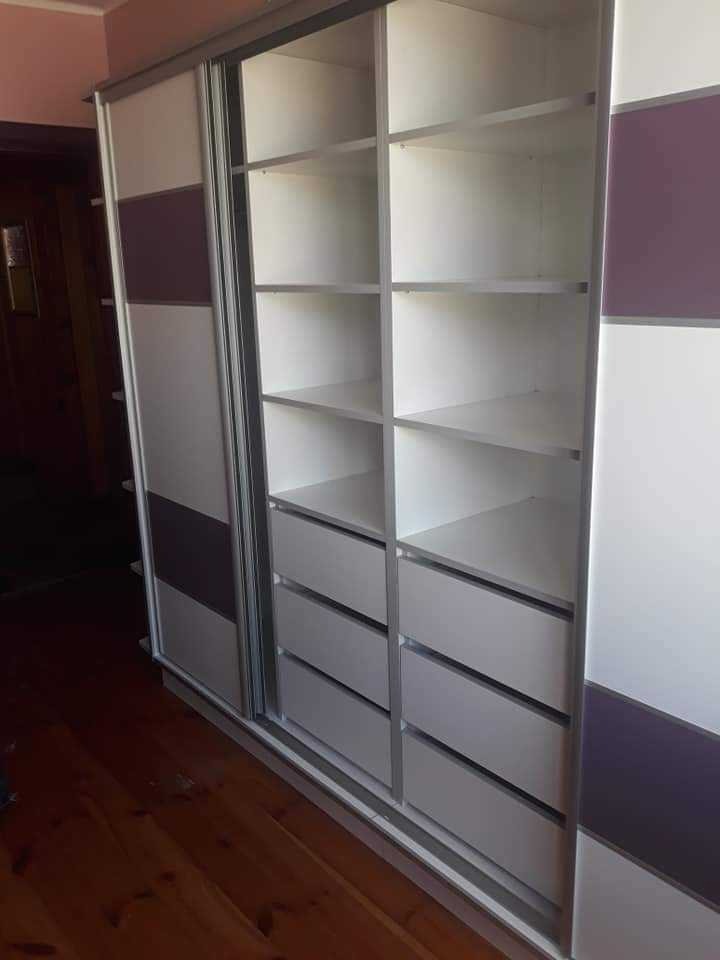 Biała szafa przesuwna z fioletowymi akcentami, widoczne półki i szuflady, drewniana podłoga, wnętrze domu.