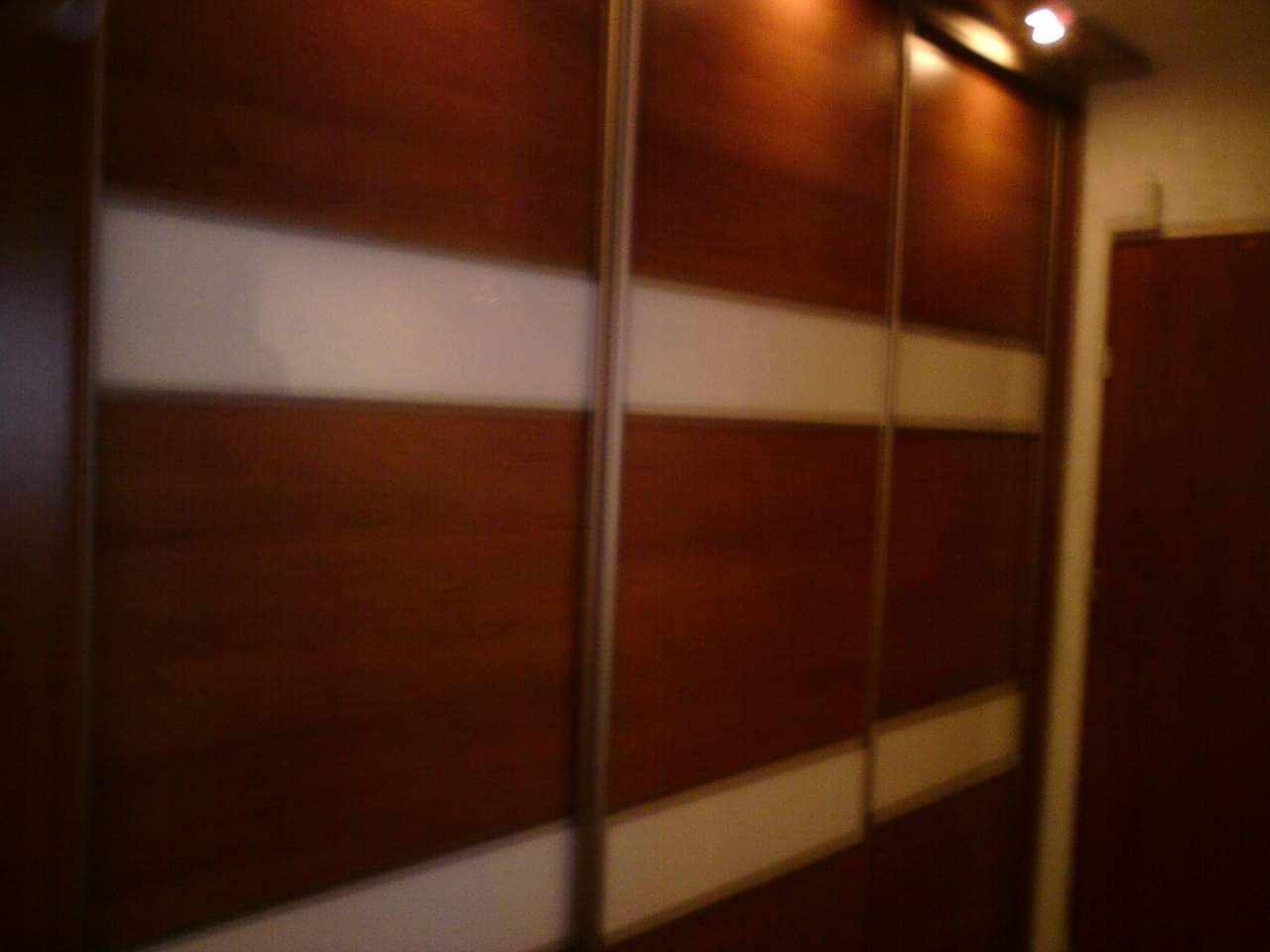 Wielodrzwiowa szafa wnękowa z przesuwnymi drzwiami, wykończona drewnem w ciepłym, brązowym odcieniu i poziomymi pasami z matowego szkła, oświetlona punktowo od góry.