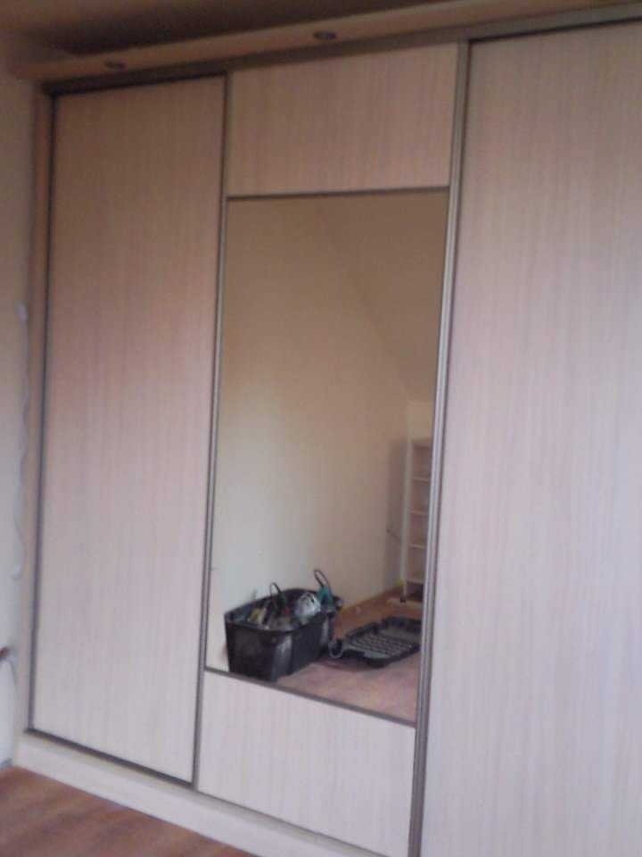 Wbudowana szafa z przesuwnymi drzwiami, wykończona jasnym drewnem i lustrem, w odbiciu widoczny pojemnik z narzędziami i drabina.