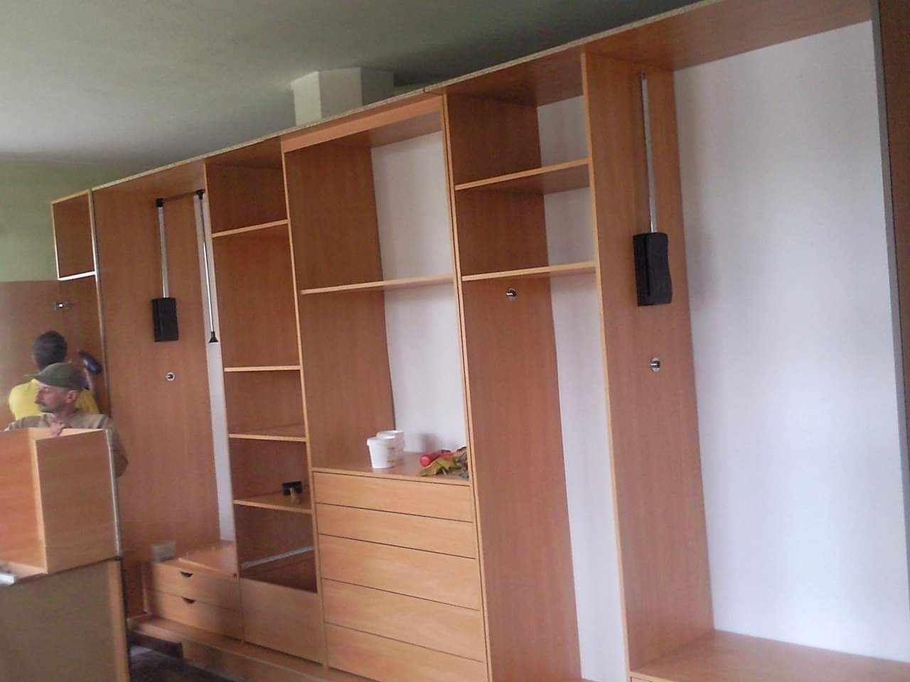 Montaż modułowej szafy na wymiar z jasnego drewna, widoczne wysuwane wieszaki, półki i szuflady, pracownik w tle.