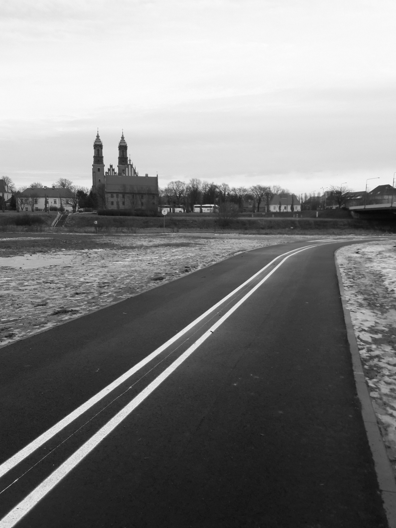 Czarno-białe zdjęcie ścieżki rowerowej z podwójną linią, prowadzącej w kierunku gotyckiego kościoła z dwiema wieżami w tle, brzeg rzeki z resztkami śniegu po bokach ścieżki.
