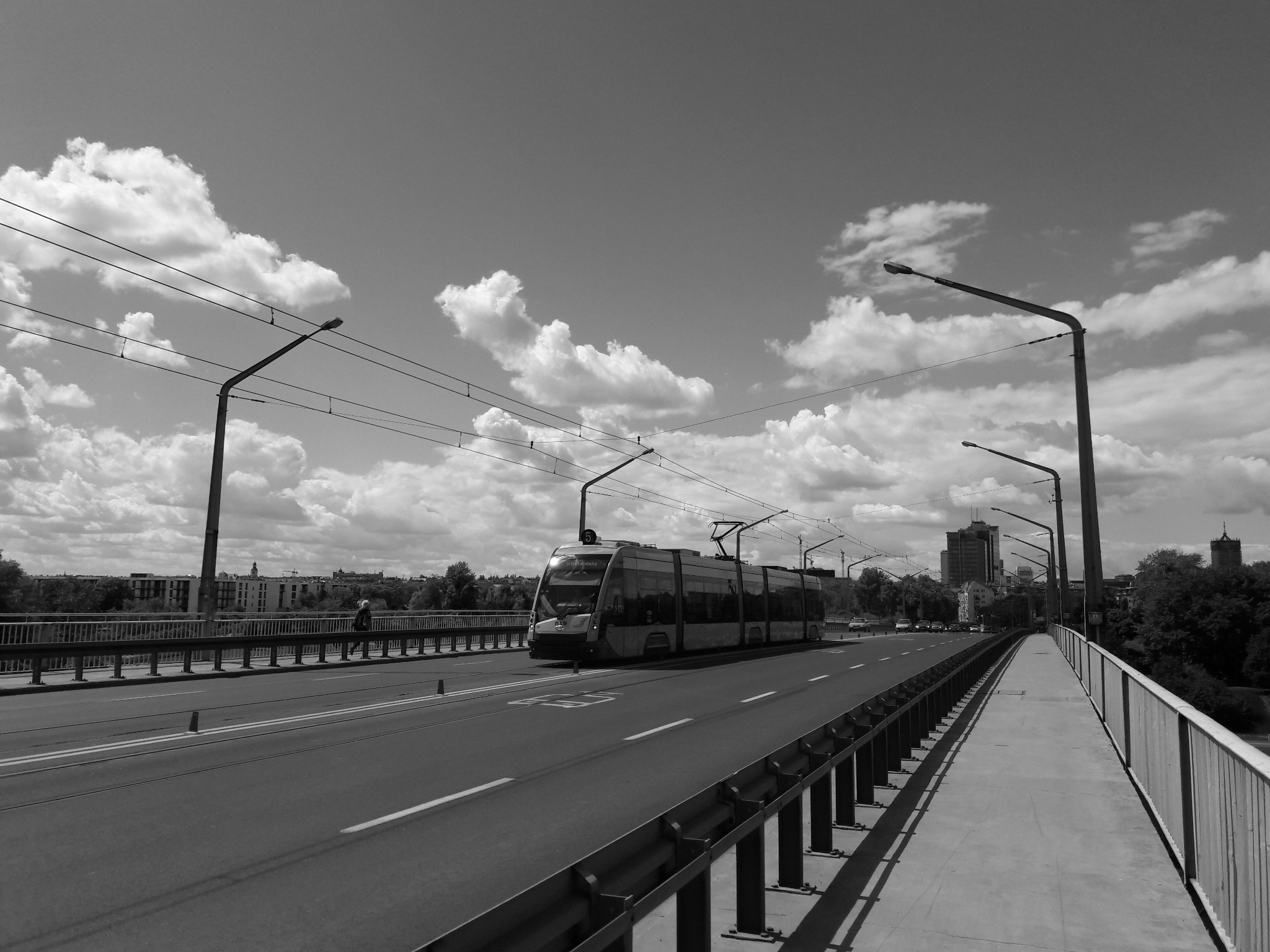 Czarno-białe zdjęcie tramwaju jadącego po moście w słoneczny dzień, z widocznymi chmurami na niebie i elementami miejskiej zabudowy w tle.