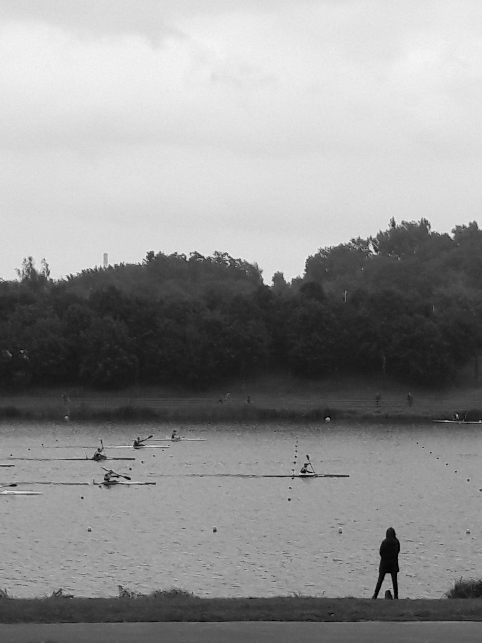 Czarno-białe zdjęcie z pochmurnego dnia nad jeziorem, na którym widać kajakarzy podczas treningu i samotną osobę stojącą na brzegu