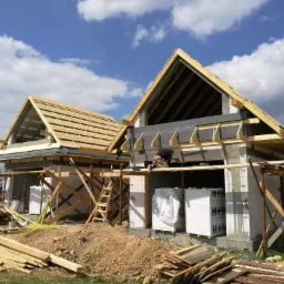 Budowa domu jednorodzinnego w stanie surowym otwartym: widoczne ściany z białego betonu komórkowego, drewniana konstrukcja dachu z deskowaniem oraz pracownik na rusztowaniu.