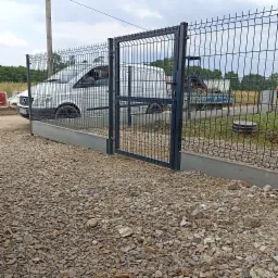 Ciemnoszara brama panelowa z zamkiem i ogrodzenie panelowe na betonowej podmurówce, z widocznym białym samochodem dostawczym i przyczepą w tle, teren wysypany drobnym kamieniem.