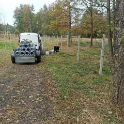 Przygotowania do montażu ogrodzenia: biały van z przyczepką pełną rolek siatki ogrodzeniowej stoi obok rzędu betonowych słupków w terenie z drzewami i trawą.