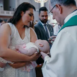 Ksiądz polewa głowę niemowlęcia wodą święconą podczas ceremonii w kościele, matka trzyma dziecko w ramionach, w tle widoczni goście.