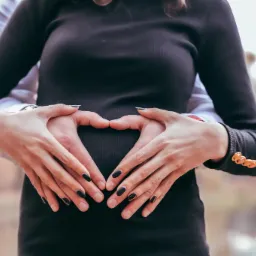 Przyszła mama w czarnej sukience, dłonie jej i partnera ułożone w kształt serca na brzuchu, w tle rozmyty krajobraz.