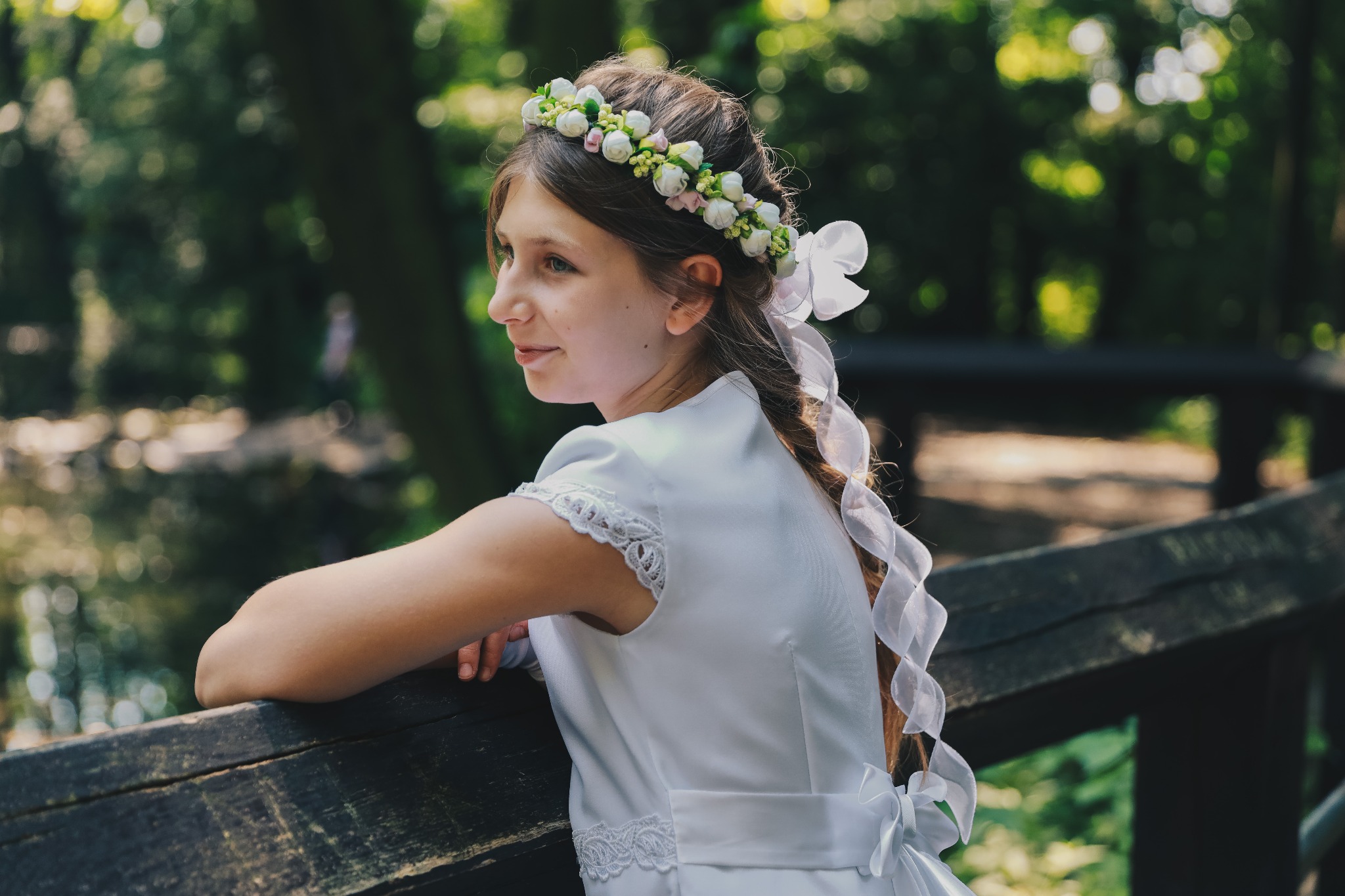 Dziewczynka w białej sukience z wiankiem na głowie, oparta o drewnianą barierkę, patrzy w dal, w tle rozmyte drzewa.
