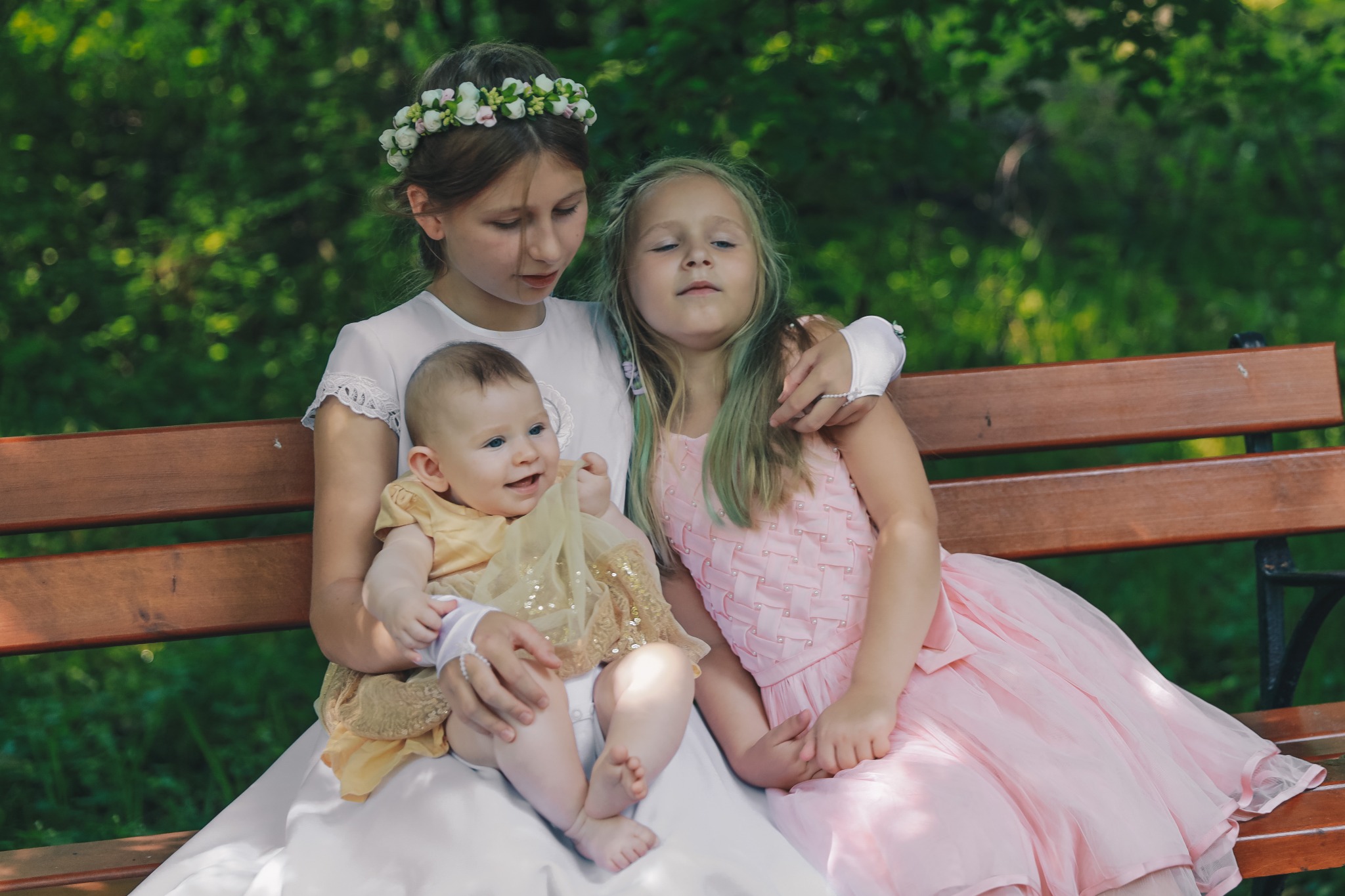Dwie dziewczynki w sukienkach, jedna z wiankiem na głowie, siedzą na ławce w parku, trzymając uśmiechnięte niemowlę ubrane w złotą sukienkę.