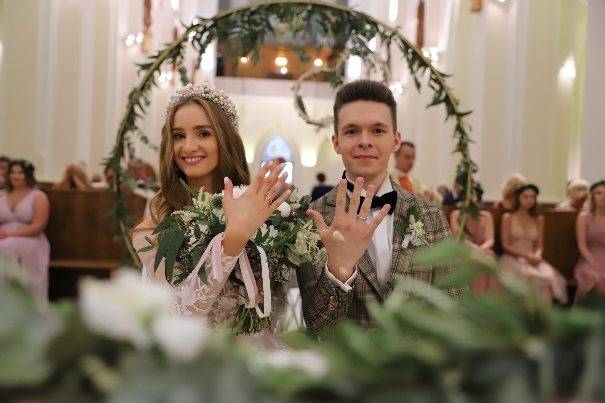 Uśmiechnięta para młoda w kościele prezentuje obrączki, panna młoda w koronie z pereł trzyma bukiet, w tle goście i dekoracja roślinna.