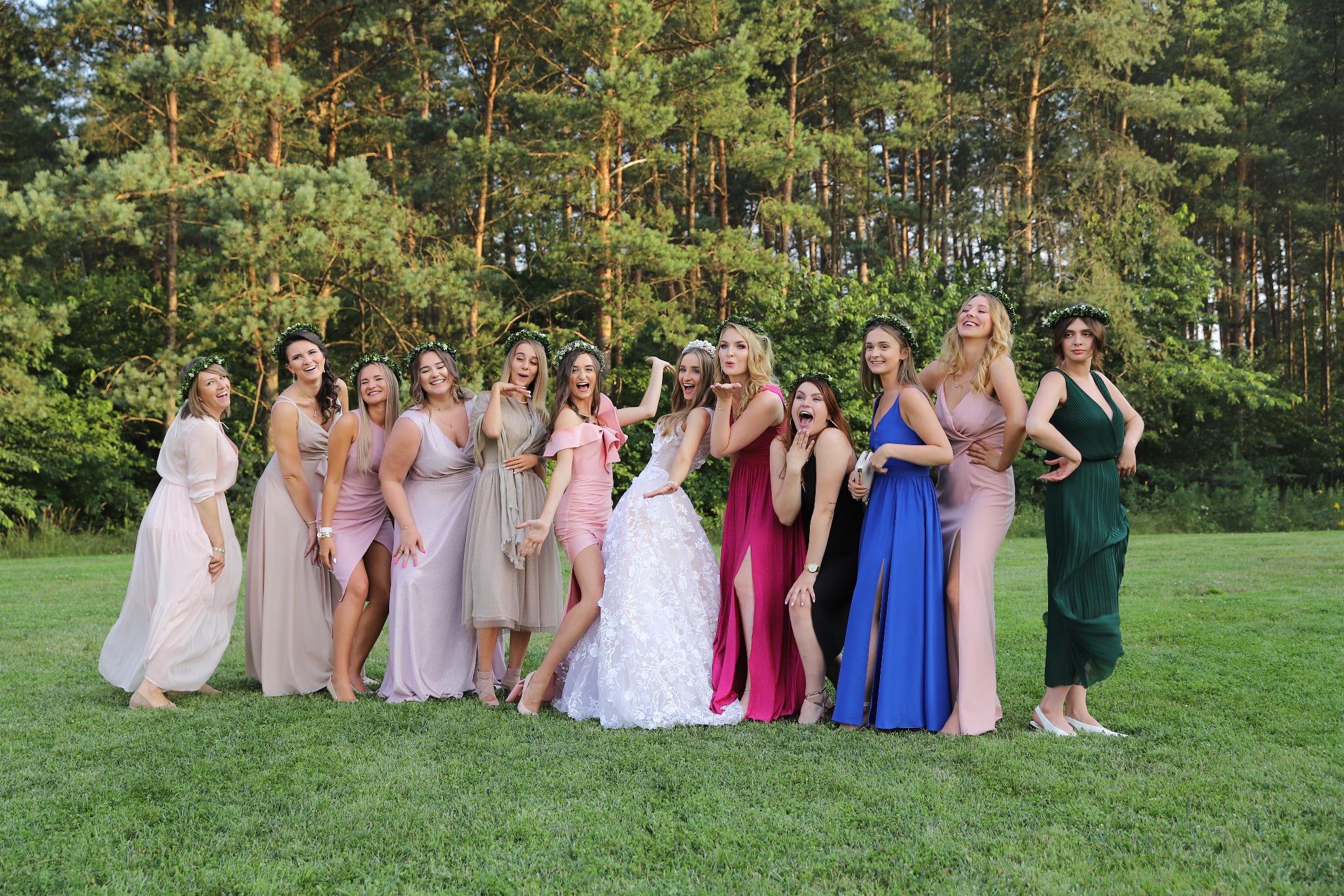 Grupa kobiet w różnorodnych sukienkach, w tym panna młoda w koronkowej sukni, pozuje na tle lasu, nosząc wianki na głowach, wyrażając radość i świętowanie.
