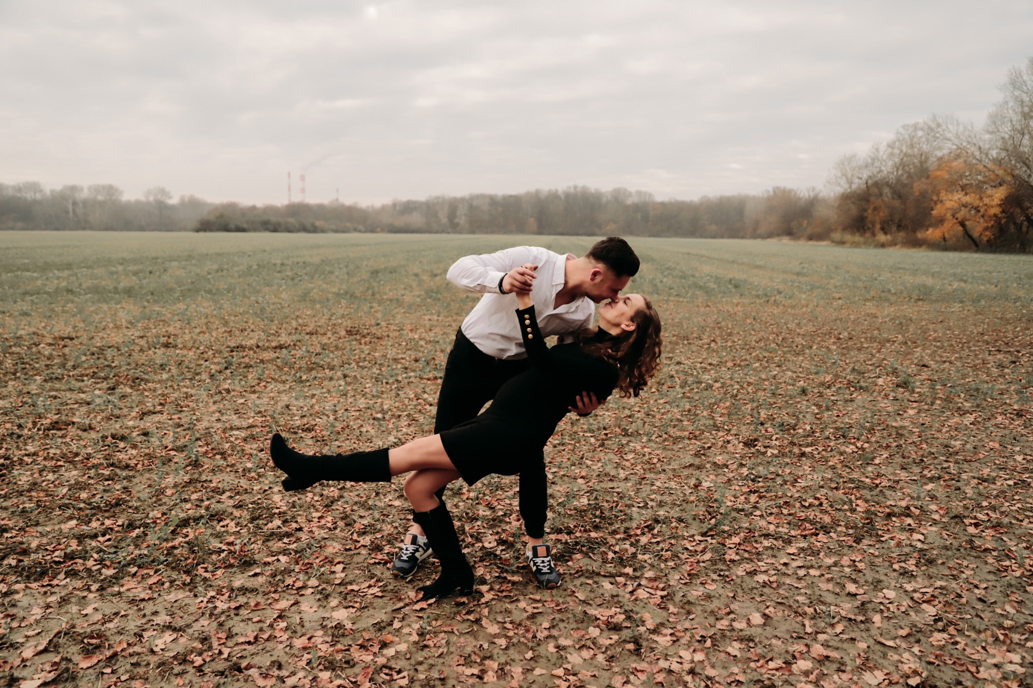 Para narzeczonych w jesiennej scenerii: mężczyzna w białej koszuli i kobieta w czarnej sukience tańczą na polu usłanym liśćmi.