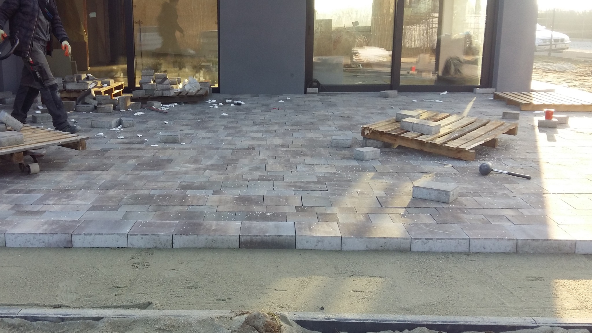 Fragment układanej nawierzchni z kostki brukowej w odcieniach szarości, widoczne palety z kostką, gumowy młotek i pracownik w trakcie robót ziemnych.