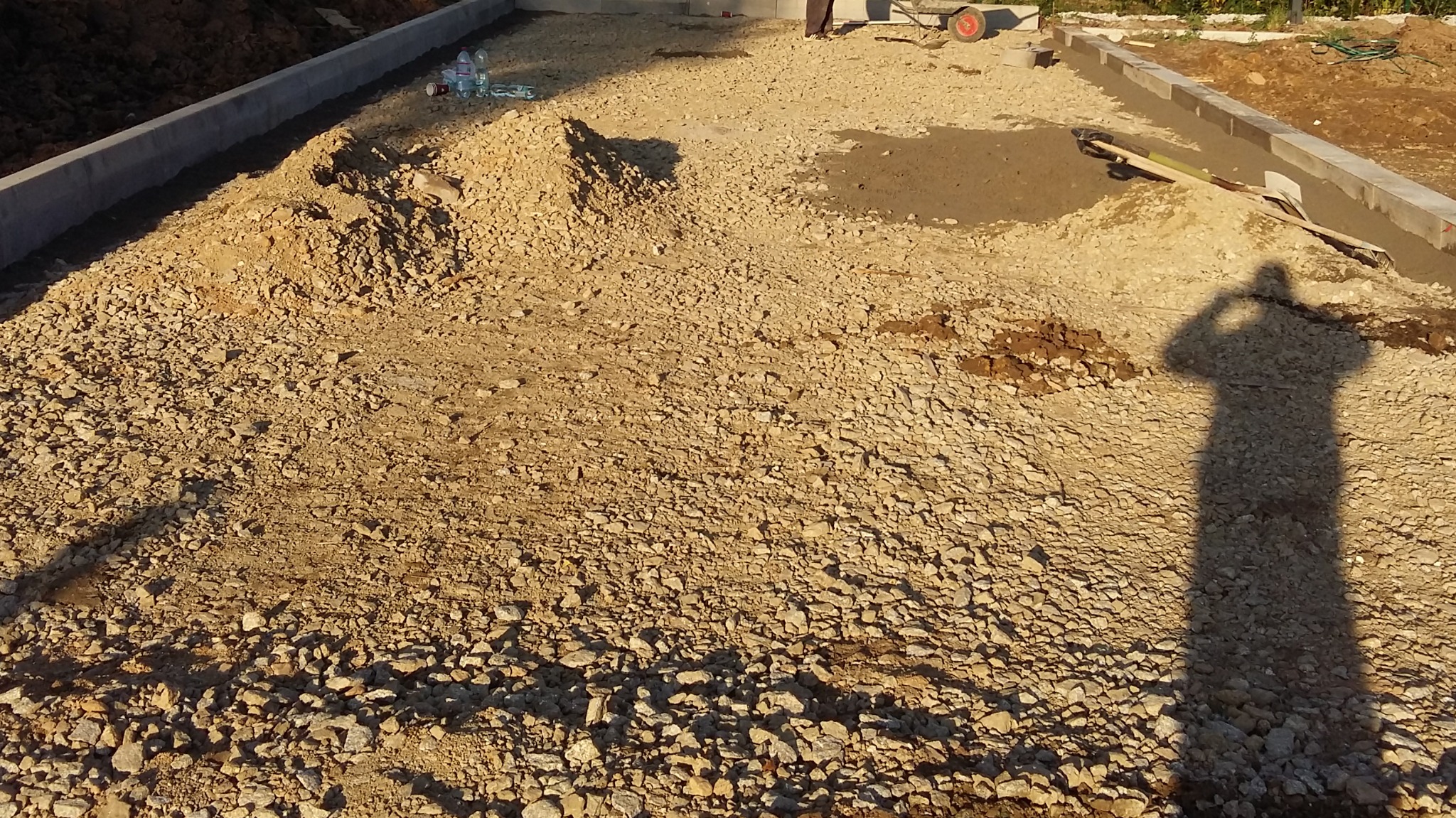 Przygotowanie podłoża pod kostkę: wysypany tłuczeń, obrzeża betonowe, narzędzia i cień osoby fotografującej.