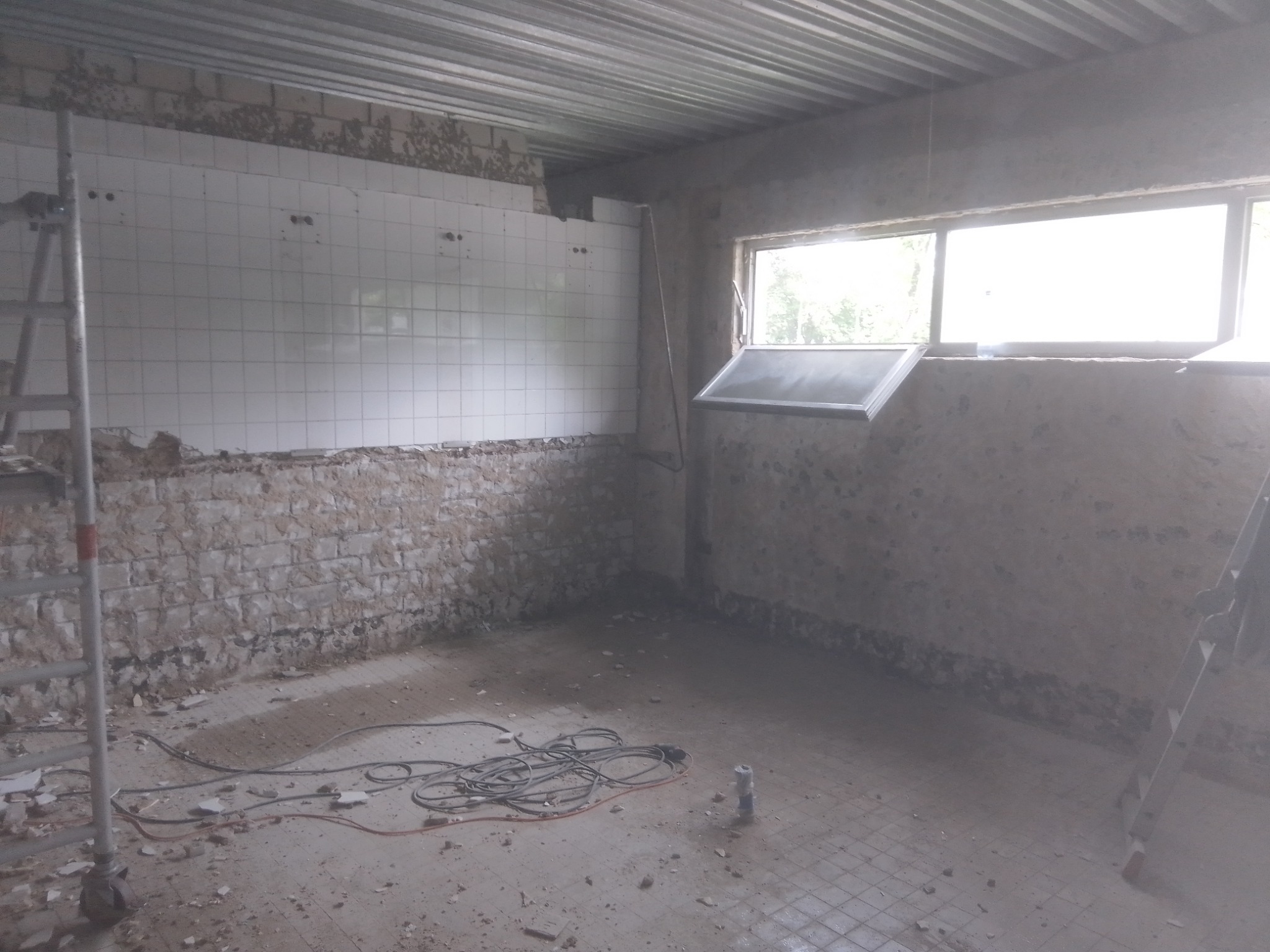 Wnętrze pomieszczenia w trakcie remontu: częściowo usunięte płytki ścienne, odsłonięta cegła, gruz na podłodze z widocznymi przewodami elektrycznymi i drabiną opartą o ścianę.