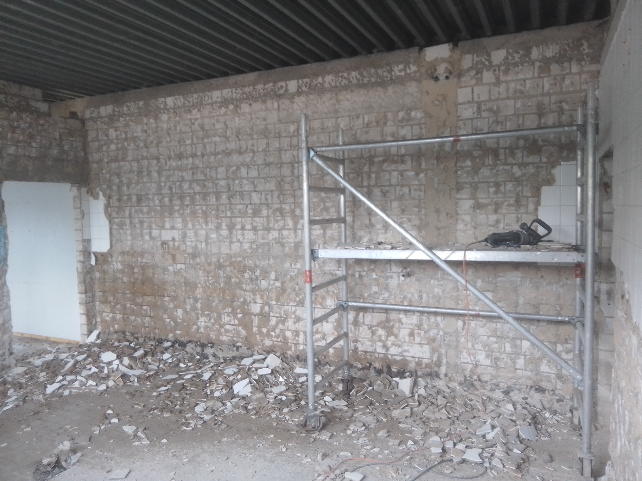 Wnętrze w trakcie demontażu płytek, widoczne rusztowanie z wiertarką udarową na górnej platformie oraz gruz na podłodze.