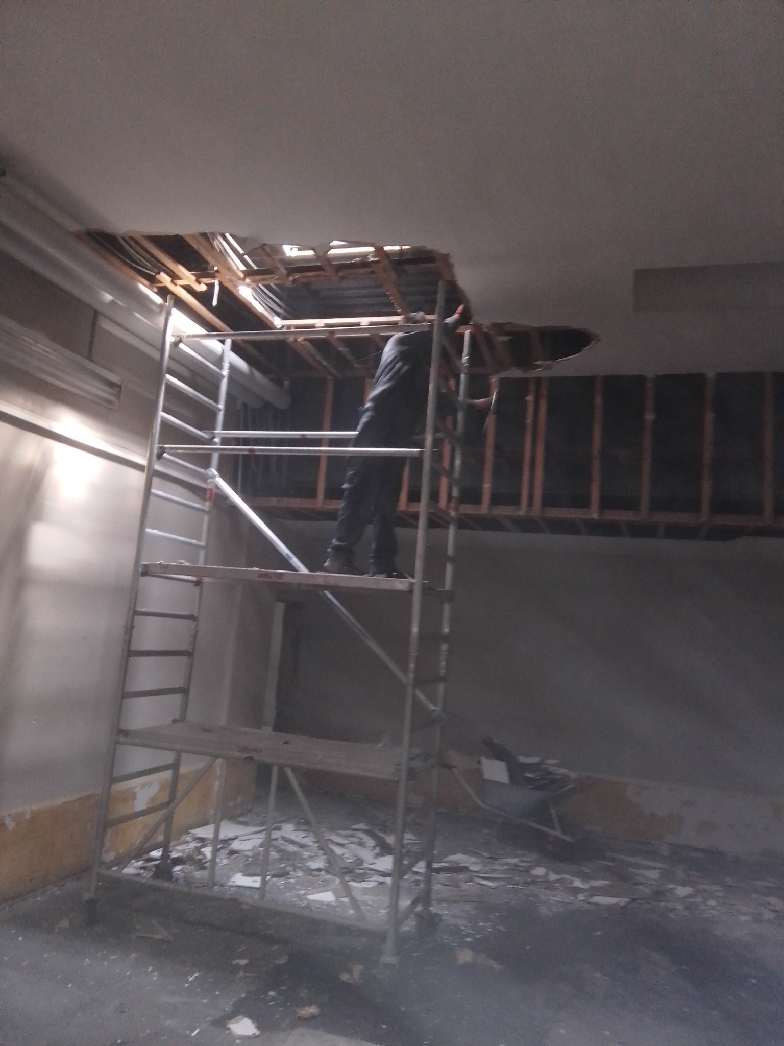 Pracownik na rusztowaniu usuwa fragment sufitu, widoczne drewniane elementy konstrukcyjne i gruz na podłodze.