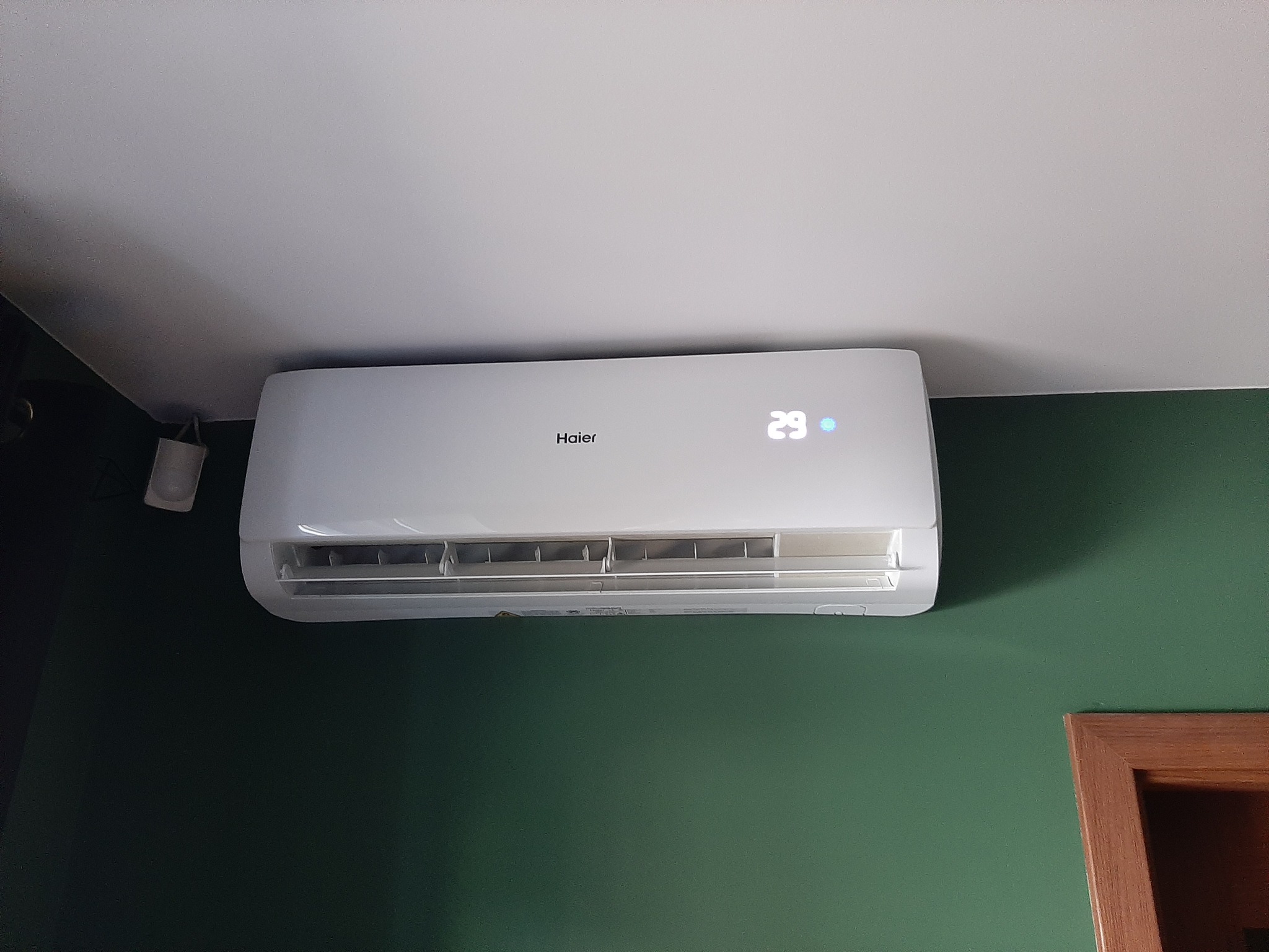 Biały klimatyzator Haier zamontowany na zielonej ścianie w pomieszczeniu, z widocznym wyświetlaczem temperatury ustawionym na 29 stopni.